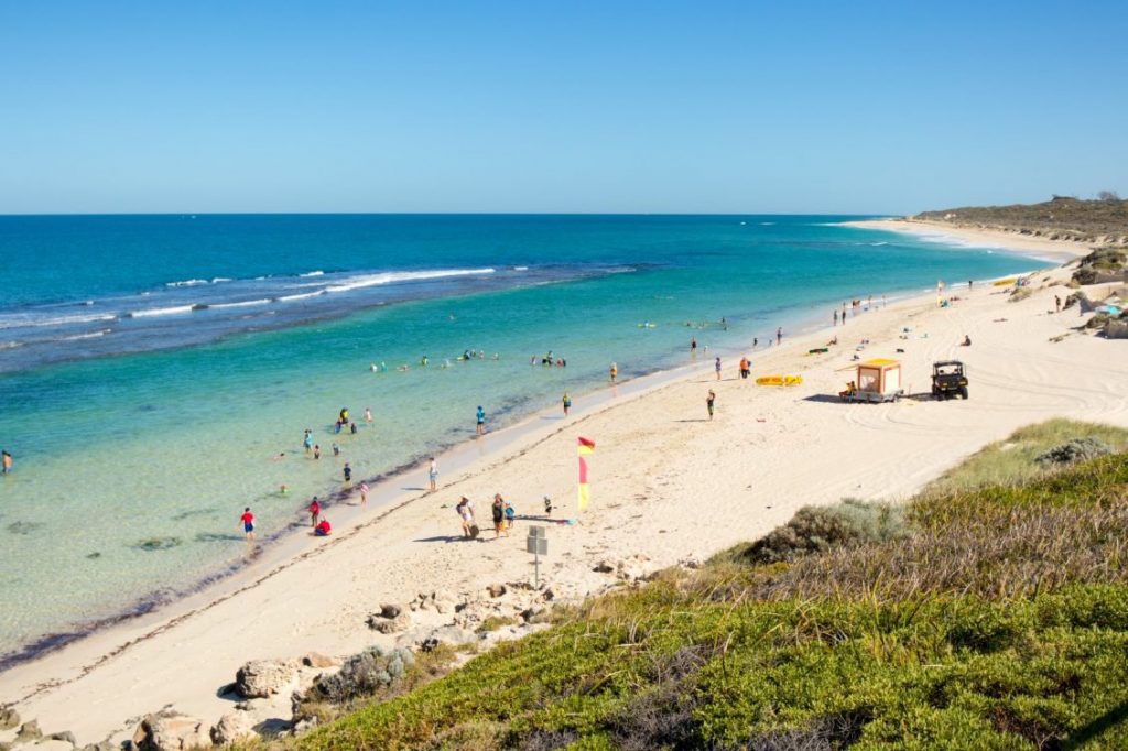 A Beach With People Swimming And Others On The Shore, Featuring Two Rocks Known As Atlantist.